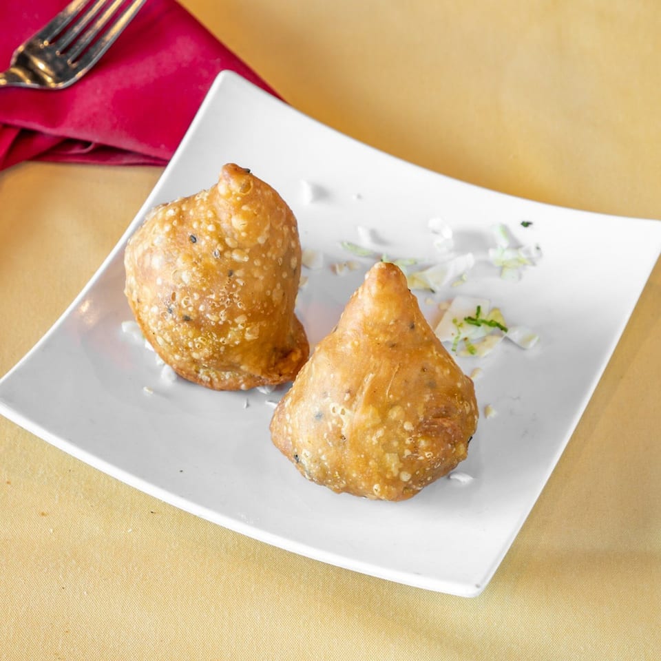 Two golden-brown Indian samosas on a white plate with shredded cabbage, a red napkin, and fork.